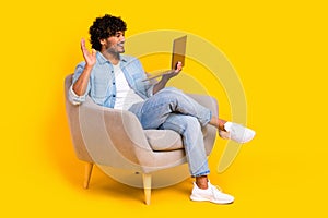 Full length photo of friendly man wear jeans shirt sit in armchair waving hand at laptop on video call isolated on