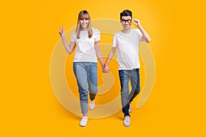 Full length photo of funny sweet young couple wear white t-shirt spectacles holding hands arms v-sign isolated yellow