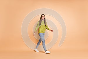 Full length photo of girl walk go look camera wear green sweater sneakers jeans isolated beige color background