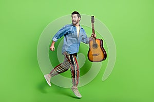 Full length photo of good mood excited guy wear denim jacket playing guitar running jumping high isolated green color