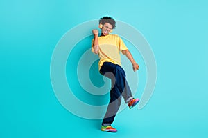 Full length photo of good mood funky guy dressed yellow t-shirt in headphones clenching fist isolated on turquoise color