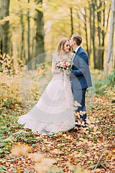 Full-length photo of the groom kissing the bride in the forehead in the autumn forest.