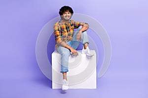 Full length photo of handsome nice man dressed plaid shirt denim pants sit on white cube platform  on purple