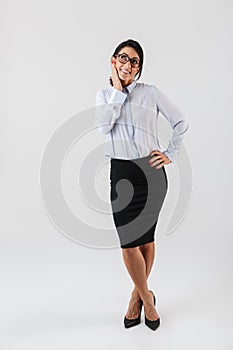 Full length photo of happy businesswoman wearing eyeglasses standing in the office, isolated over white background