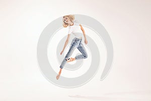 Full length photo of happy, smiling, young blonde girl in casual jeans and white t-shirt jumping, dancing over light studio