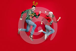 Full length photo of jumping couple excited by x-mas prices hurry buy costumes wear ugly ornament jumpers isolated red