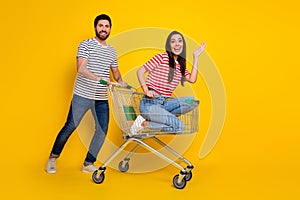Full length photo of lovely couple wear t-shirt sit in cart ride to empty space on black friday isolated on yellow color