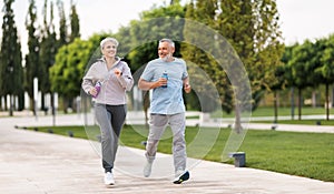Full-length photo of lovely joyful retirees couple jogging outside in city park photo