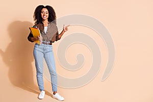 Full length photo of nice girl wear brown blouse holding copybook indicating at promo empty space isolated on pastel