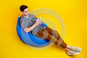 Full length photo of nice man dressed print shirt brown trousers look at smartphone lay on bean bag isolated on yellow