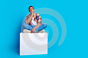 Full length photo of nice young man sit cube look empty space wear shirt isolated on blue color background