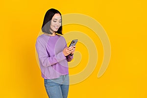 Full length photo of optimistic curious woman wear purple shirt jeans shoes look at phone typing post isolated on yellow
