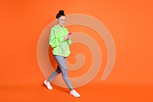 Full length photo portrait of woman texting holding phone in two hands walking isolated on vivid orange colored