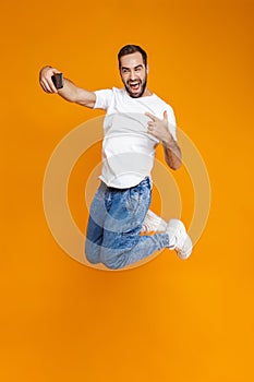 Full length photo of positive guy 30s in casual wear laughing and taking selfie on cell phone, isolated over yellow background