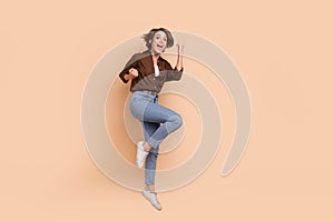 Full length photo of pretty excited girl dressed brown shirt jumping high rising fists empty space isolated beige color