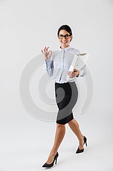 Full length photo of pretty female secretary wearing eyeglasses holding paper folder in the office, isolated over white background