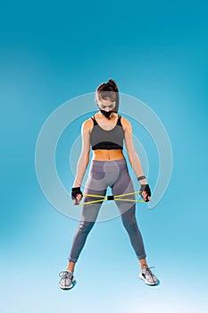 A full-length photo of a pumped-up girl in a protective black mask and with a resitance band in her hands. Looks down on