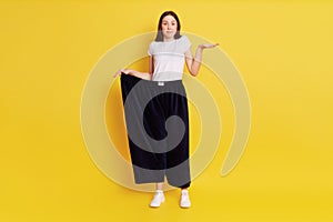Full length photo of puzzled astonish female wearing too big blag pants, stretching palm aside, looking at camera, being surprised