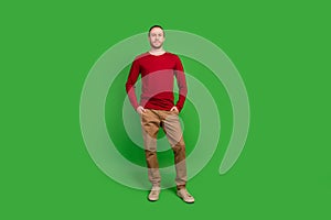Full length photo of serious handsome guy wear red sweatshirt brown trousers keep palms in pockets isolated on green