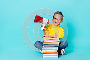 Full length photo of small cheerful boy sitting floor pile stack book hand hold loudspeaker isolated on cyan color