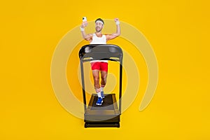 Full length photo of strong funny guy sportswear running treadmill rising fist tacking selfie device isolated yellow