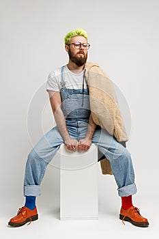 Full length photo of stylish model man wearing trendy clothing and red shoes, sitting on white cube, looking away with dreamy