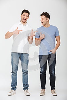 Full length photo of two handsome men 30s wearing casual t-shirt and jeans smiling and gesturing fingers on each other, isolated