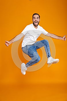 Full length photo of unshaved guy in t-shirt and jeans jumping and having fun,  over yellow background