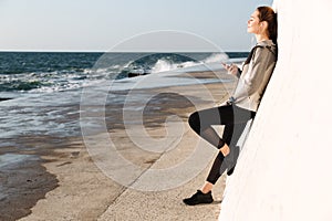 Full-length photo of young charming woman in sport wear listening to music with closed eyes, standing near white wall, seaside