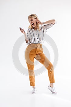 Full length photo of young woman singing and holding mobile phone while listening to music via earpod