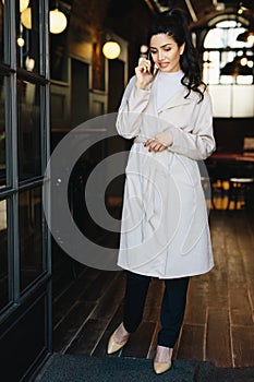 Full length portait of elegant businesswoman wearing white overcoat, stylish shoes and black trousers standing in cafe calling he