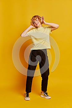 Full-length portrait of active young red-haired girl dancing isolated on yellow studio background. Human emotions