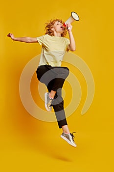 Full-length portrait of active young red-haired girl jumping with megaphone isolated on yellow studio background. Human