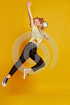 Full-length portrait of active young red-haired girl jumping with megaphone isolated on yellow studio background. Human