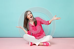 Full length portrait of angry young woman in rose shirt blouse, white pants sitting on floor isolated on bright pink