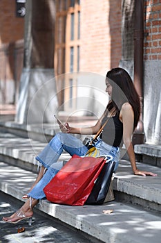 Portrait of Asian woman sitting on stairs with shopping bags and using smart phone.