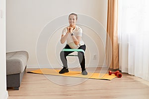 Full length portrait of athletic woman wearing white t shirt and black leggins, doing squats with resistance band on knees,