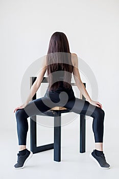 Full-length portrait of the attractive charming brunette in sportsuit sitting on the chair with the long hair on her
