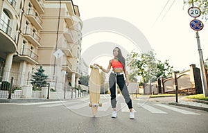 Full length portrait of attractive girl in stylish street clothes, standing on street with longboard in hands, looking into camera