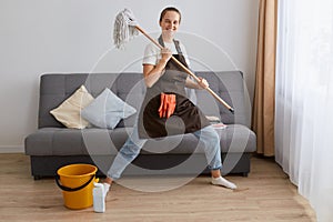 Full length portrait of attractive happy smiling woman wearing jeans and brown apron holding mop in hands and dancing while