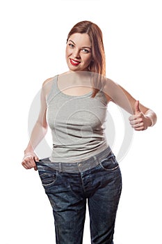 Full-length portrait of attractive slim young smiling woman in big jeans showing successful weight loss with her thumb up, isolate