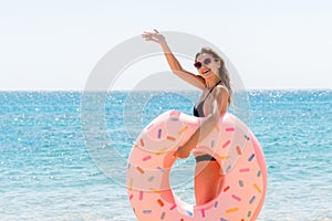 Full length portrait of attractive woman holding doughnut inflatable ring and waving her hand on the beach at the sea background.