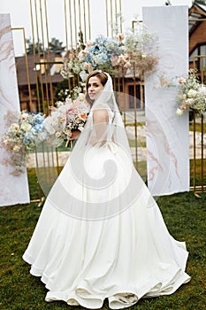 Full length portrait of attractive young bride in a white wedding dress and veil holding a big bouquet of white and pink