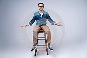 Full length portrait of an attractive young man in jeans shirt sitting on the chair over grey background.