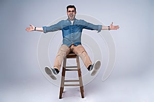 Full length portrait of an attractive young man in jeans shirt sitting on the chair over grey background.