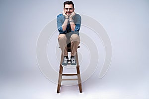 Full length portrait of an attractive young man in jeans shirt sitting on the chair over grey background.