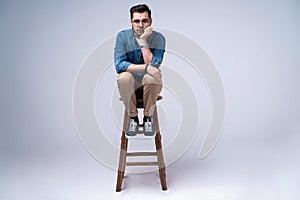 Full length portrait of an attractive young man in jeans shirt sitting on the chair over grey background.