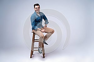 Full length portrait of an attractive young man in jeans shirt sitting on the chair over grey background.