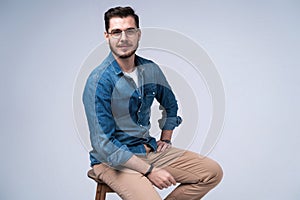 Full length portrait of an attractive young man in jeans shirt sitting on the chair over grey background.