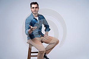 Full length portrait of an attractive young man in jeans shirt sitting on the chair over grey background.